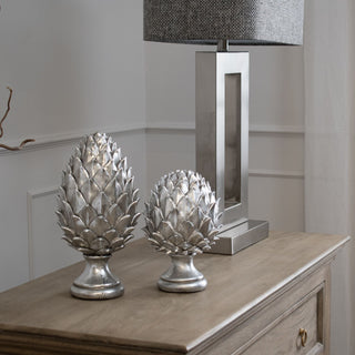 Two silver ceramic pinecone ornaments, large and small, displayed on a wooden side table next to a metal lamp.
