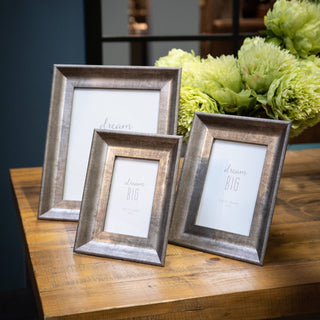 Three distressed silver convex frames displayed together on a rustic wooden table with green flowers.