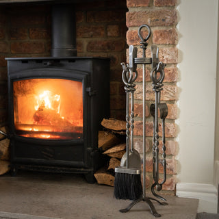 Fireplace tool set displayed next to a lit wood-burning stove in a brick hearth setting.