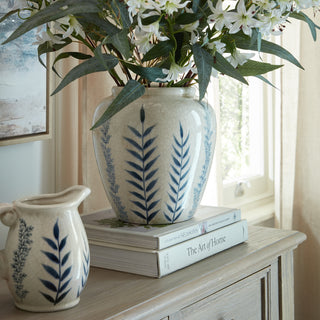Cream crackle-glazed vase with blue leaf design, filled with white flowers, displayed on books on a wooden cabinet next to a matching jug.