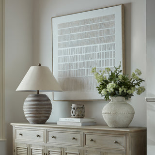 Textured abstract canvas art displayed above a wooden sideboard styled with a large lamp and a vase of white flowers.
