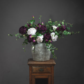 Aged stone effect vase holding purple and white faux flowers, sitting on a rustic wooden pedestal.