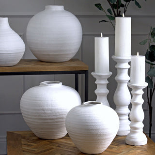 Styled scene showing three white column candle holders of varying heights alongside textured white ceramic vases.