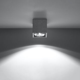 Concrete ceiling light fixture illuminated, showing the focused downward beam against a grey wall.