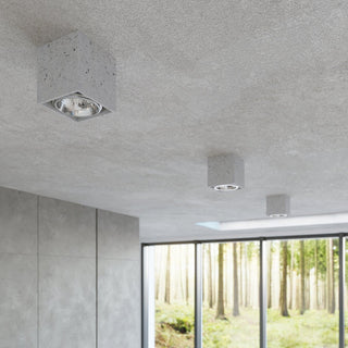 Three concrete cubic lights mounted on a textured ceiling in a minimalist room with large windows overlooking a forest.