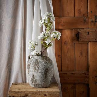 Blossom stem in a rustic grey urn on a wooden crate, styled against a sheer white curtain and panelled door.