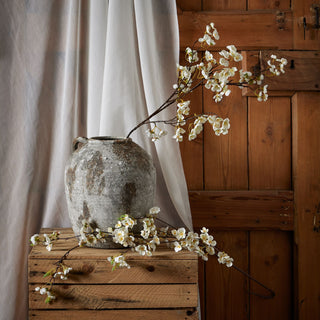 Styling shot of artificial white blossom in a large urn on wooden crates, set against sheer fabric and rustic wood.