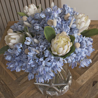 White bunny tails mixed into a large blue and white floral arrangement in a vase on a wooden table.