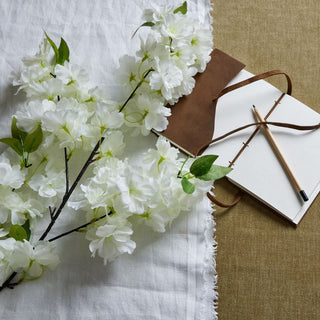 Close-up of the white artificial cherry blossom stem styled on white fabric next to a brown leather notebook and pencil.