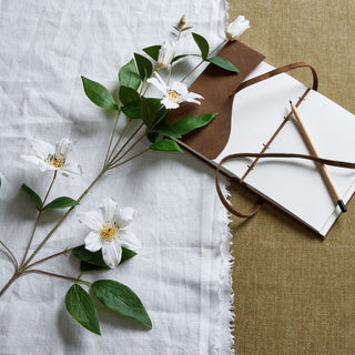 White clematis stem with green leaves laid across a white cloth, styled with a brown leather notebook and pencil.