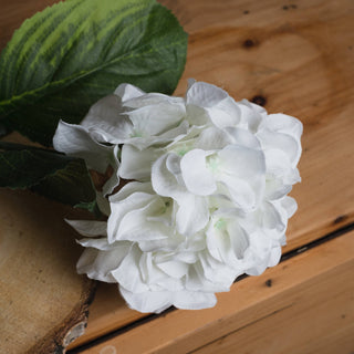 Artificial white hydrangea head shown close up, with a green leaf on rustic wooden planks.