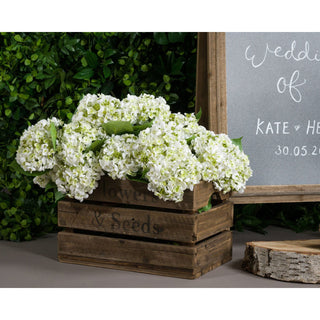 Several white hydrangea heads in a rustic wooden crate, styled with a chalkboard sign and green hedge.