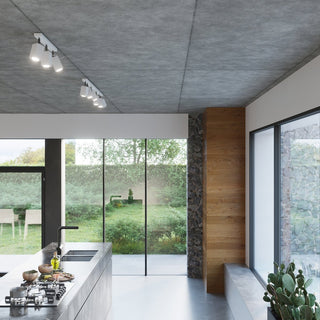 White track spotlights mounted on a raw concrete ceiling in a modern kitchen with wood panelling and glass walls overlooking a garden.