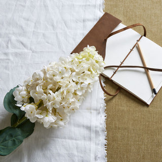 White spear hydrangea stem on white linen and brown fabric, styled with an open notebook and pencil.