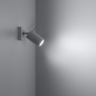 White cylindrical spotlight mounted in a corner, angled to cast a bright beam of light onto the adjacent wall.